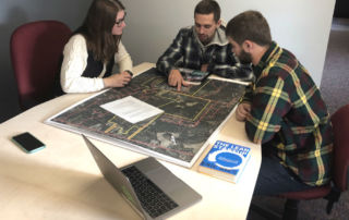 Three people are seated around a table in MABLab, examining a large map, with a laptop, notebook, and a book titled "The Lean Startup" on the table. This dynamic scene reflects the spirit of entrepreneurism prevalent in their on-campus internship. Saint Joseph's College of Maine