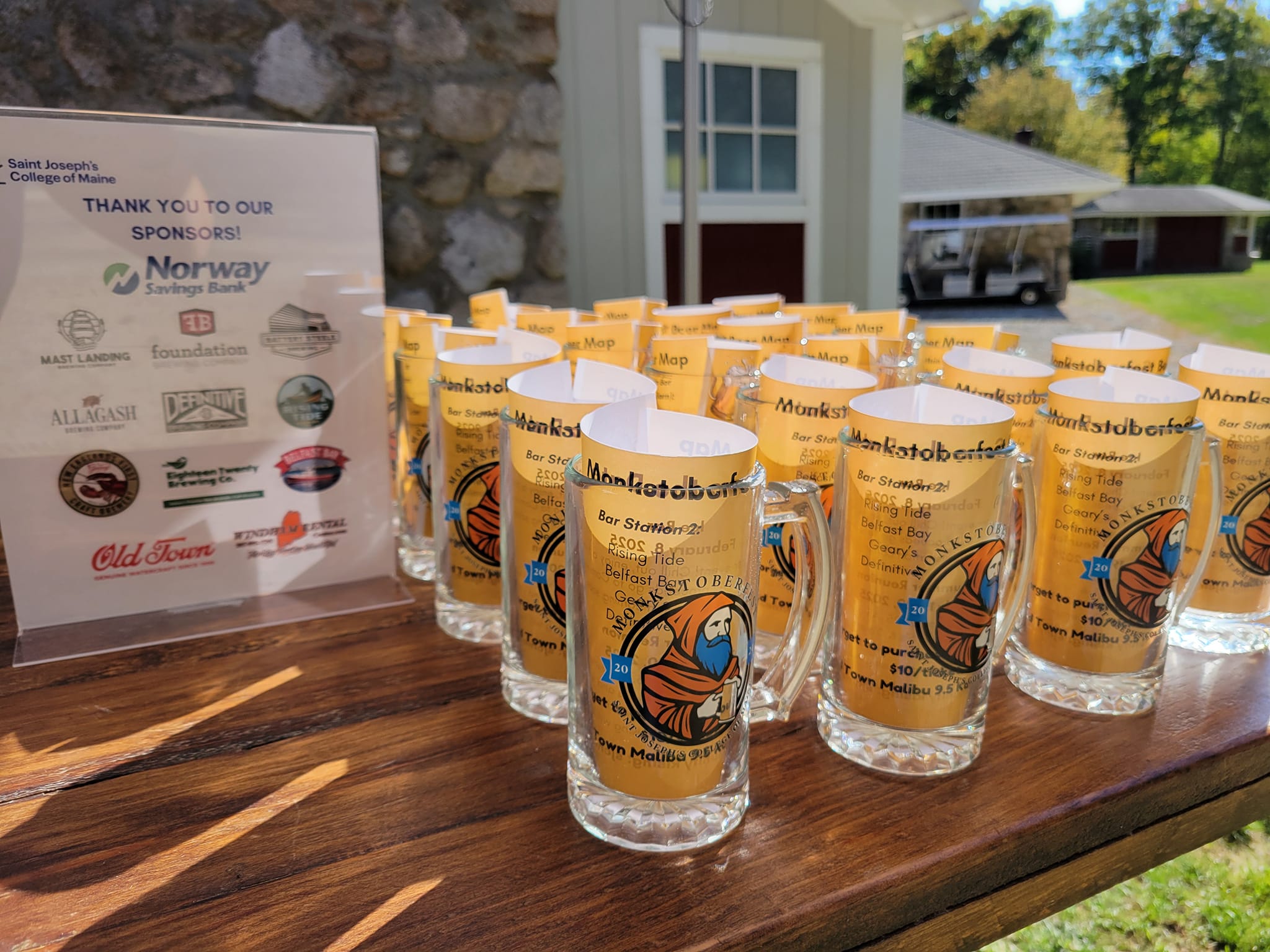 A table displays multiple beer glasses with event logos. A sponsorship sign is visible on the left. The setting appears to be outdoors with buildings and greenery in the background. Saint Joseph's College of Maine