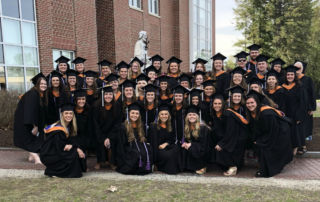 A group of graduates wearing caps and gowns pose together outside Saint Joseph’s College of Maine, with large windows and a statue in the background, celebrating not just their degrees but also the impressive NCLEX pass rate achieved through the college's esteemed BSN programs. Saint Joseph's College of Maine