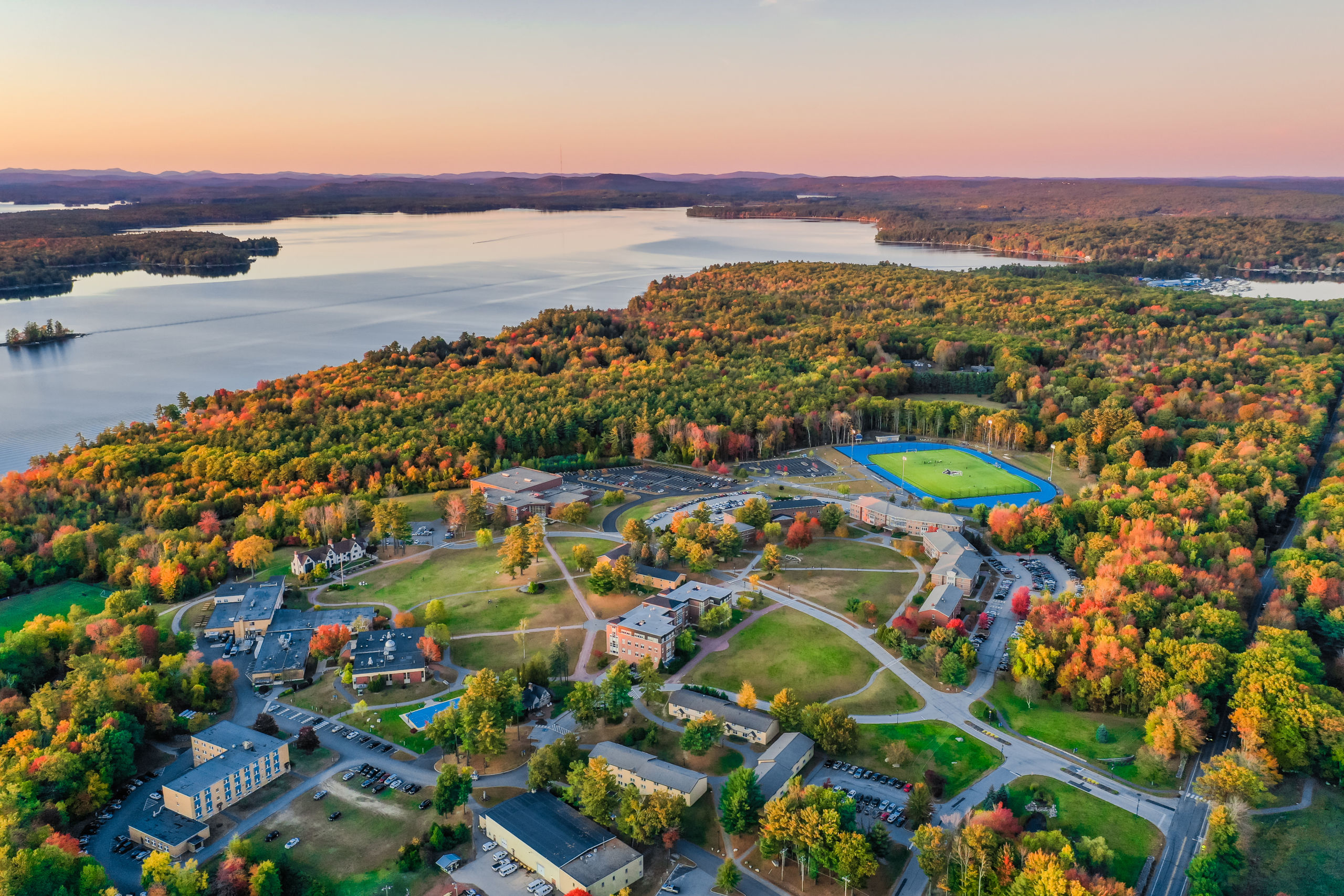 Fall Drone 2019 Aerial view of a campus surrounded by autumnal trees near a large lake at sunset. Streets and buildings are visible, with a sports field in the background. Saint Joseph's College of Maine