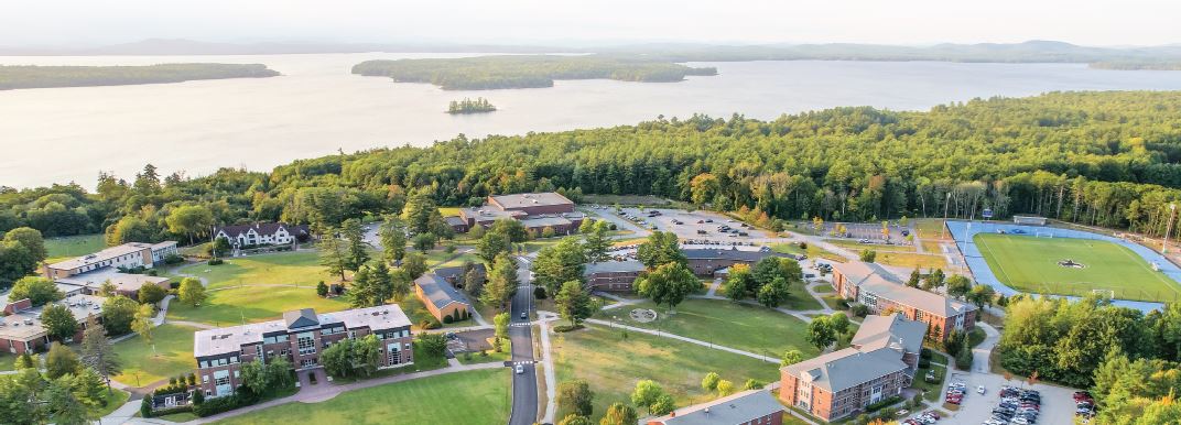 Aerial view of a college campus surrounded by trees, with several buildings, a sports field, parking lots, and a large lake in the background. Saint Joseph's College of Maine