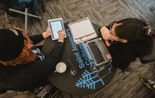 Two people sit at a round table with "Saint Joseph's College" written on it. One holds a tablet and stylus, the other a laptop and notebook. A coffee cup and earphones are also visible on the table, perhaps celebrating making the Dean's List for fall 2023. Saint Joseph's College of Maine