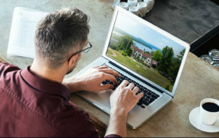 A person with glasses is working on a laptop at a countertop. Next to the laptop, there is a cup of coffee and a book bearing the logo of Saint Joseph's College in Maine. Saint Joseph's College of Maine
