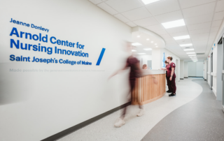 The hallway in the nursing center buzzes with activity as a sign for the Arnold Center for Nursing Innovation catches the eye. A person walks by while another stands near the reception desk, discussing opportunities for nursing students funded by a recent HRSA Grant. Saint Joseph's College of Maine