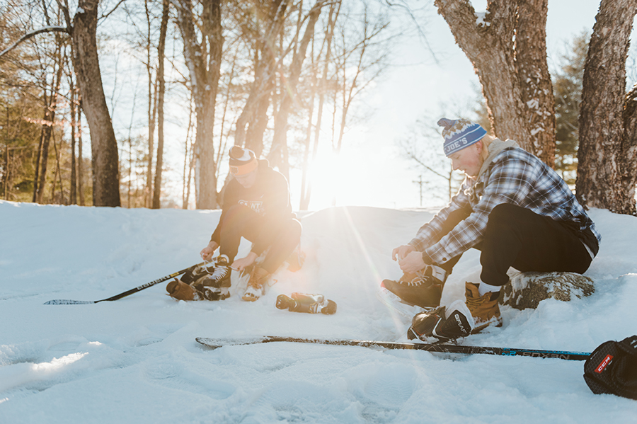 Two people sit in the snow putting on ice skates next to hockey sticks, with the sun shining between leafless trees in the background, a perfect setting for student activities. Saint Joseph's College of Maine