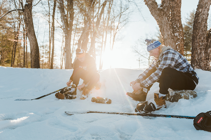 Skate Two people sit in the snow putting on ice skates next to hockey sticks, with the sun shining between leafless trees in the background, a perfect setting for student activities. Saint Joseph's College of Maine