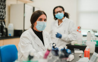 Two scientists wearing masks and lab coats are working in a laboratory. One is seated, while the other stands, both handling laboratory equipment and samples. Saint Joseph's College of Maine