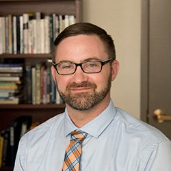 Bradley-Campbell A man with glasses and a beard wearing a light blue shirt and a striped tie stands in front of a bookshelf, likely deep in thought about criminal justice degrees in Maine. Saint Joseph's College of Maine