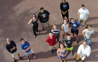 Communication Students Take To The Skies With Video-Capturing Drone. 5 A group of twelve communication students standing outdoors on a brick-paved area, looking up at the camera. They are casually dressed and appear to be smiling as a video-capturing drone hovers in the skies above them. Saint Joseph's College of Maine