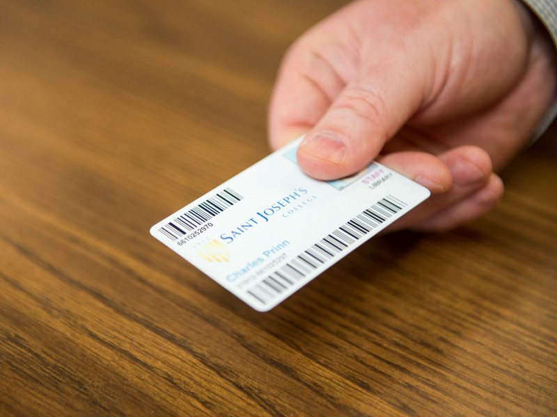 Community-Members-2 A hand holding a Saint Joseph's College ID card with barcodes and the name Charles Primn visible on the wooden table background, right next to an entrance brochure for Wellehan Library. Saint Joseph's College of Maine