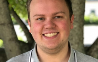 A man wearing a light gray collared shirt and flip-flops smiles at the camera, with green foliage and tree trunks in the background, hinting at an ankle deep exploration of nature. Saint Joseph's College of Maine