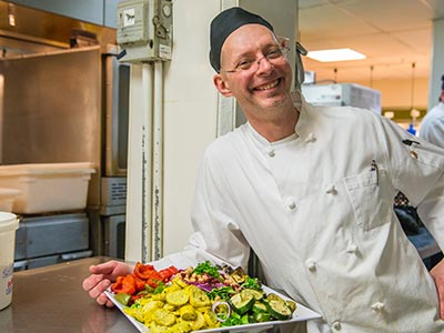 Food-Business-Incubator A chef stands in a kitchen, smiling and holding a plate of colorful, assorted vegetables, showcasing the bounty sourced through the Institute for Local Food Systems Innovation. Saint Joseph's College of Maine