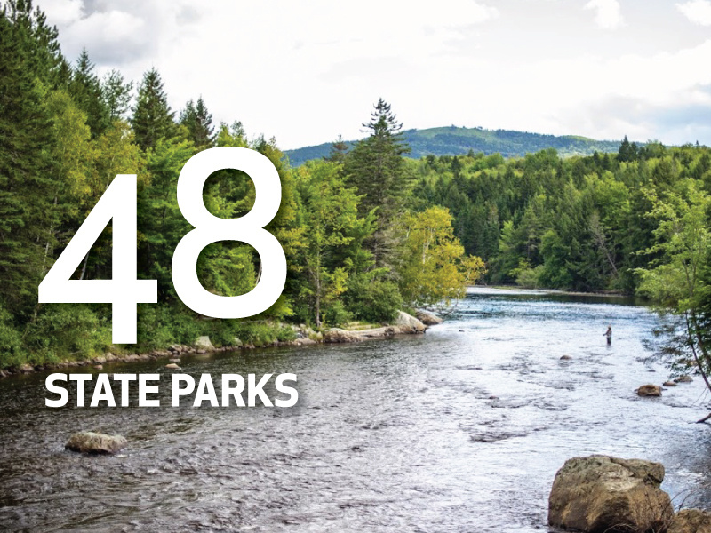 View of a river with dense trees on either side and hills in the background, featuring large text "48 State Parks" over the image. Explore Maine's scenic beauty and discover its natural wonders. Saint Joseph's College of Maine
