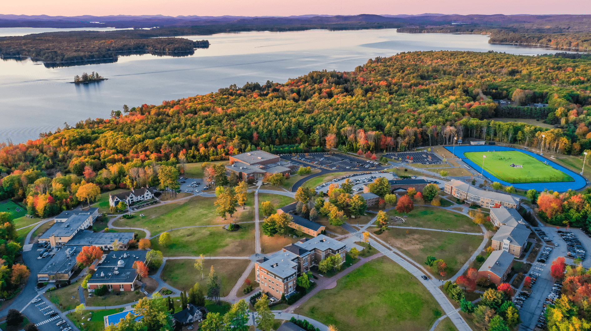 Image-3 Aerial view of a university campus in the fall, surrounded by colorful trees and situated near a large lake. Buildings, pathways, a sports field, and parking lots are visible. Saint Joseph's College of Maine