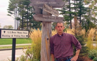 A man stands next to a wooden directional signpost with multiple arrows. The background features a sign for L.L. Bean stores amidst trees and tall grass, offering a behind the scenes glimpse into an outdoor adventurer's haven. Saint Joseph's College of Maine