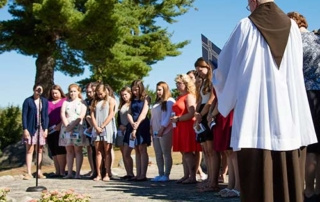 2016 Junior Education Blessing 4 A group of people stand outdoors in a semi-circle. A person in a white robe faces them, offering a Junior Education blessing near a microphone. Trees and the clear sky of 2016 serve as the backdrop. Saint Joseph's College of Maine