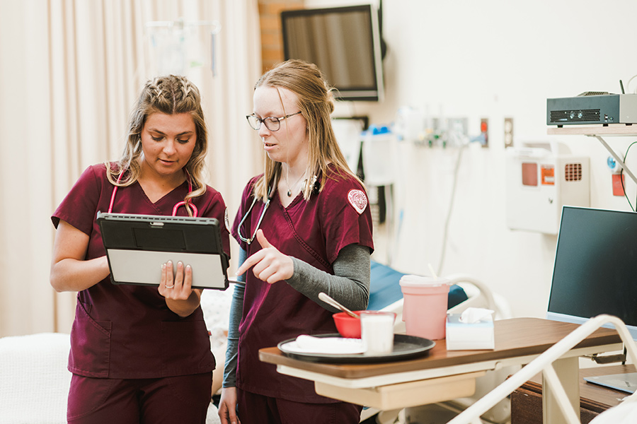 Two nurses in maroon scrubs review patient information on a tablet in a hospital room filled with medical equipment, showcasing the practical applications of online health administration programs. Saint Joseph's College of Maine