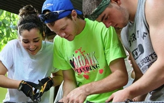 The College Celebrates 10 Years Of Service Trips To Guatemala 2 Three young adults, celebrating 10 years of college service trips in Guatemala, work together to cut and secure wires. One wears a bright green shirt and holds wire, another uses pliers, and a third wears a headband. Saint Joseph's College of Maine