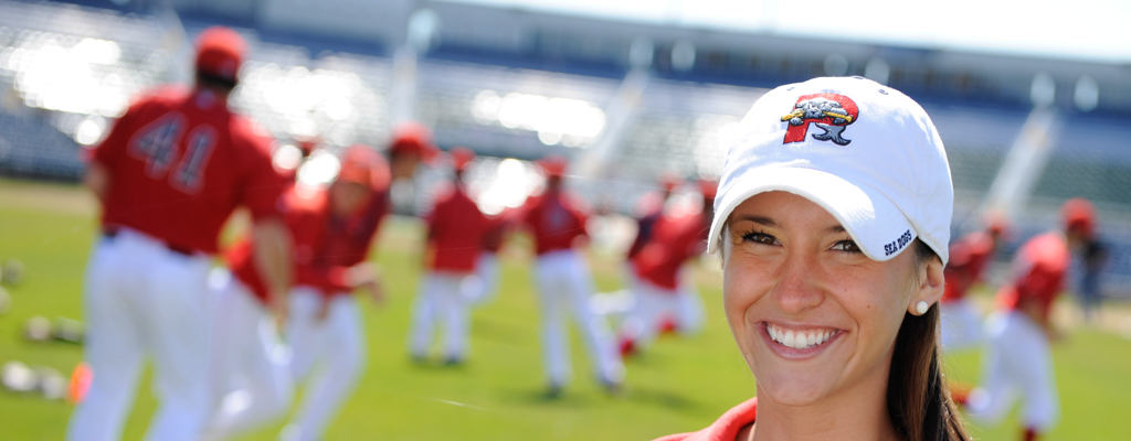A smiling woman in a white baseball cap stands in the foreground, while a group of people in red uniforms practice on a baseball field in the background. Her experience and dedication are enhanced by her sports management degree, making her well-equipped to navigate the dynamic world of athletics. Saint Joseph's College of Maine