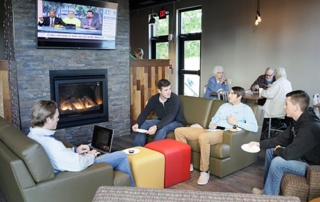 Saint Joseph’s College Opens A New Dining Experience On Campus 3 Four people sit and talk in a café lounge with a fireplace and a TV showing news in the background. Other patrons, including students from Saint Joseph’s College, are seated at tables behind them, enjoying the new dining experience on campus. Saint Joseph's College of Maine